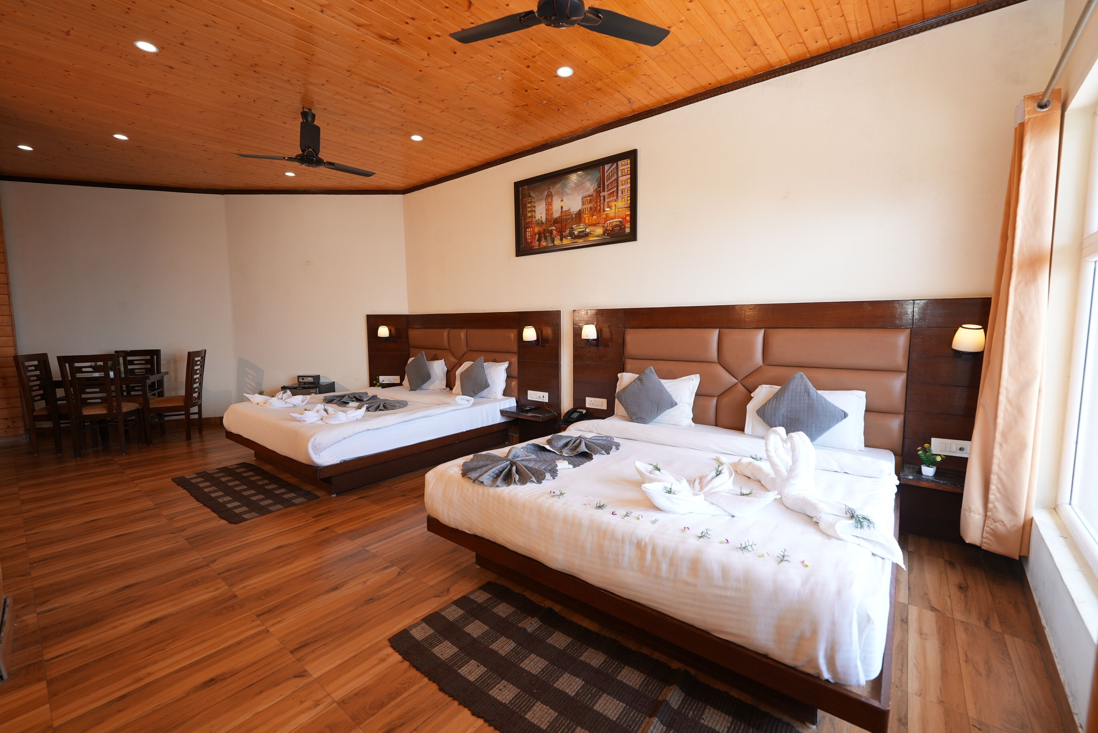 Habitación de hotel con dos camas, piso de madera, ventilador de techo, un cuadro en la pared y un comedor con sillas.
