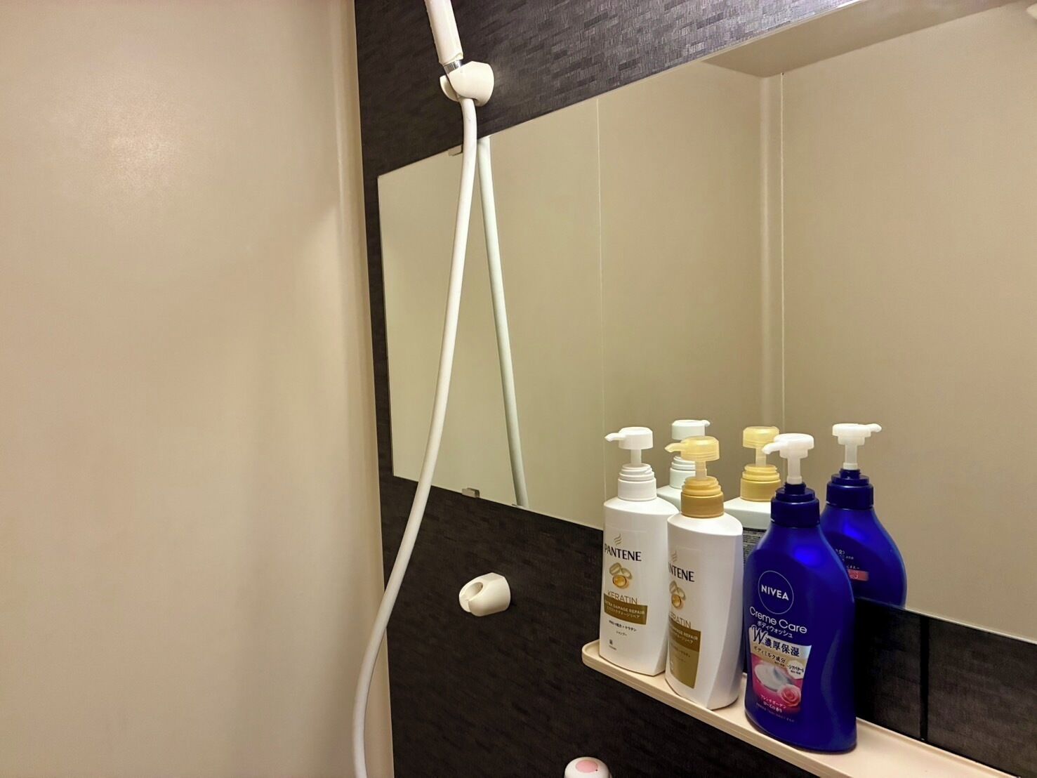 A bathroom with a mirror, a white showerhead, and shelves with various toiletries including Pantene and Nivea products.