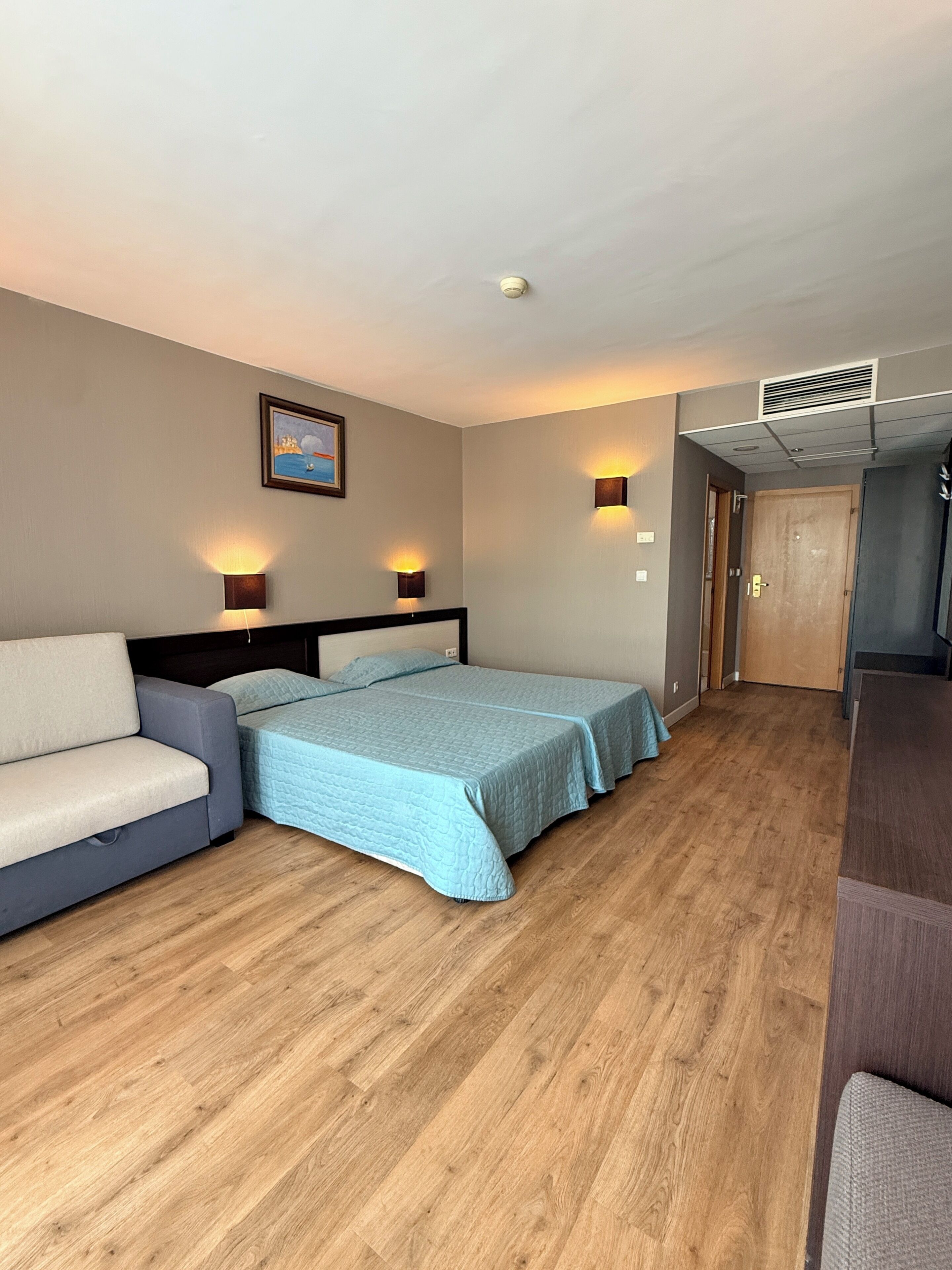 A hotel room with a bed, a sofa, a painting on the wall, and a door.
