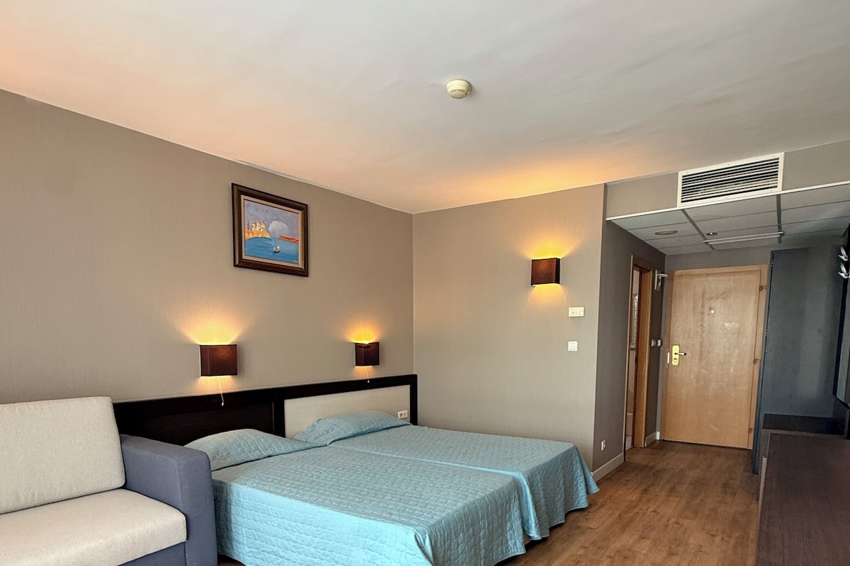 A hotel room with a bed, a sofa, a painting on the wall, and a door.