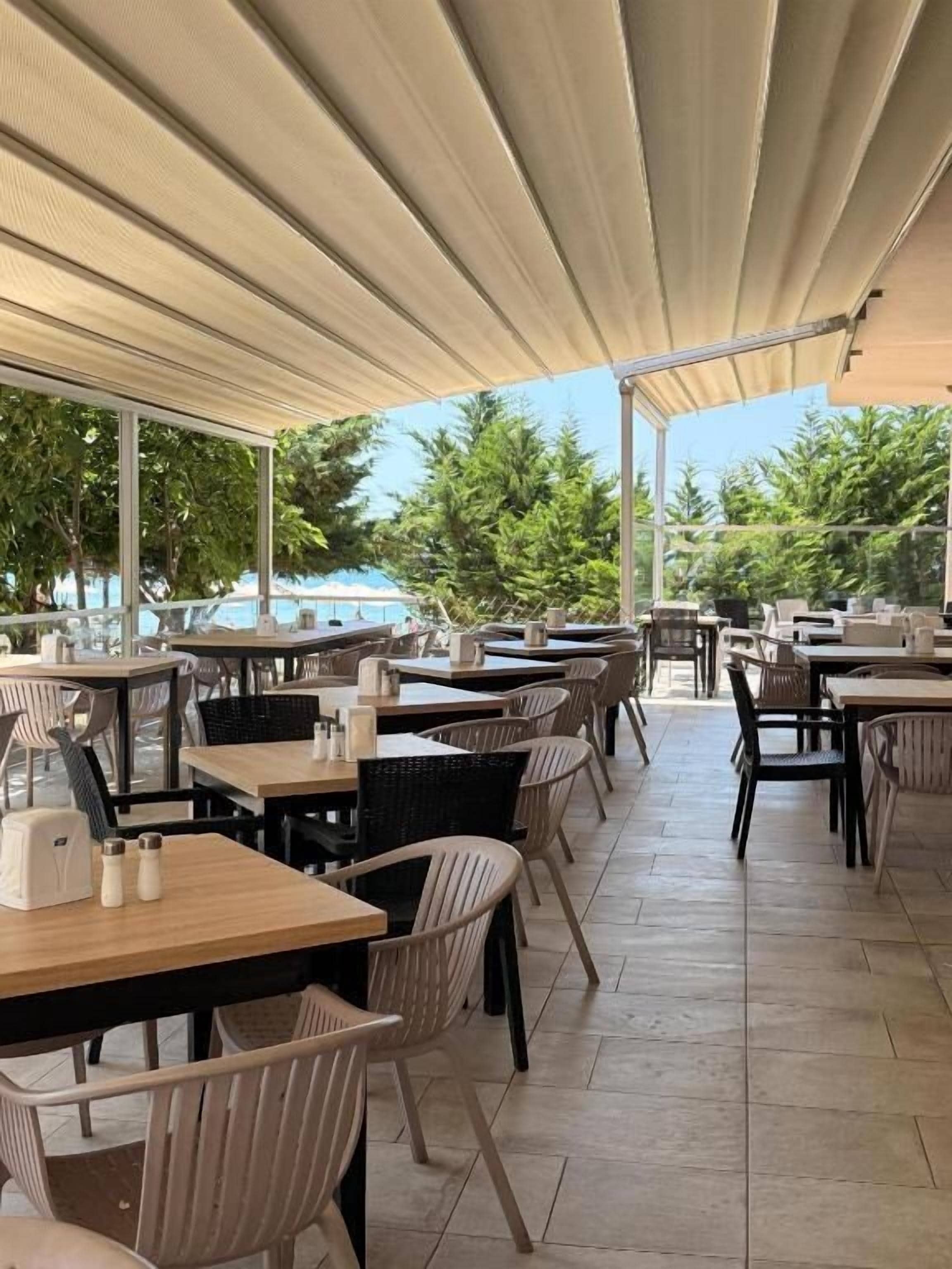 Un café al aire libre con mesas y sillas, un área techada y vistas a una piscina y zonas verdes.