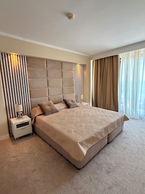 A large bed with a headboard, a bedside table with a lamp, and a window with curtains.