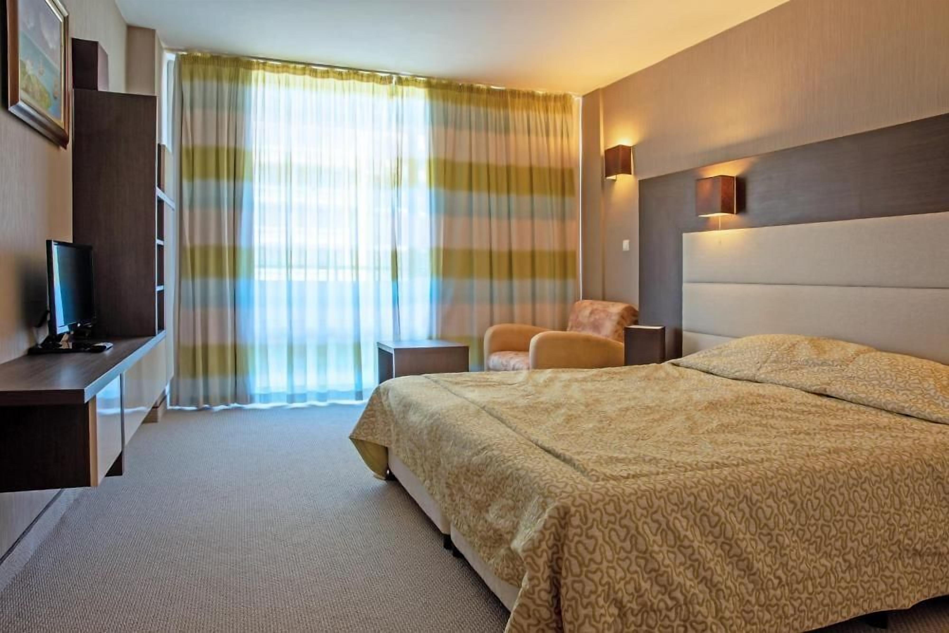 Ett hotellrum med en säng, ett skrivbord med en dator, en stol och ett fönster med gardiner.