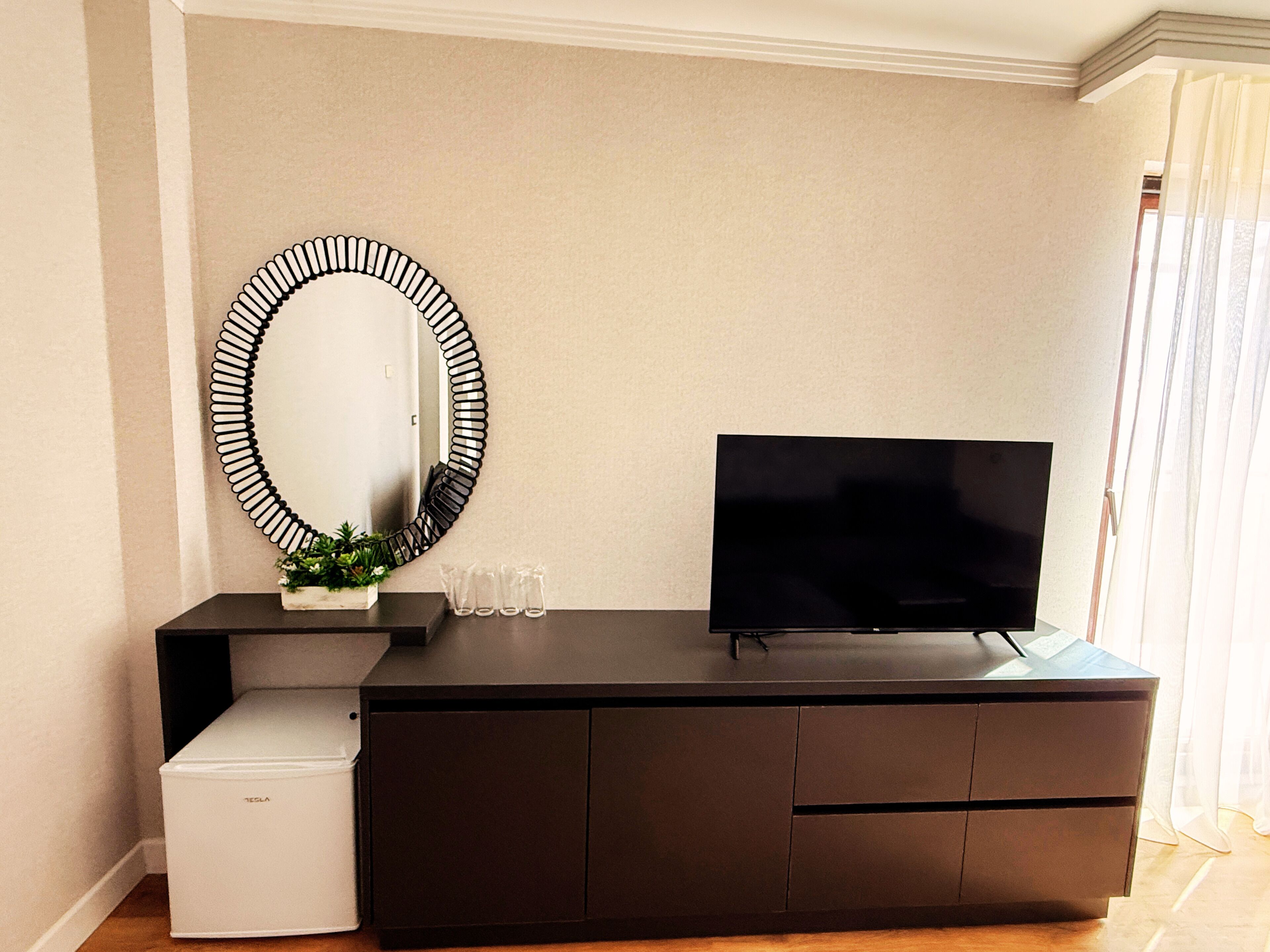 A modern living room with a flat-screen TV, a dark wooden cabinet, a white mini-fridge, and a unique black and white spiral mirror.