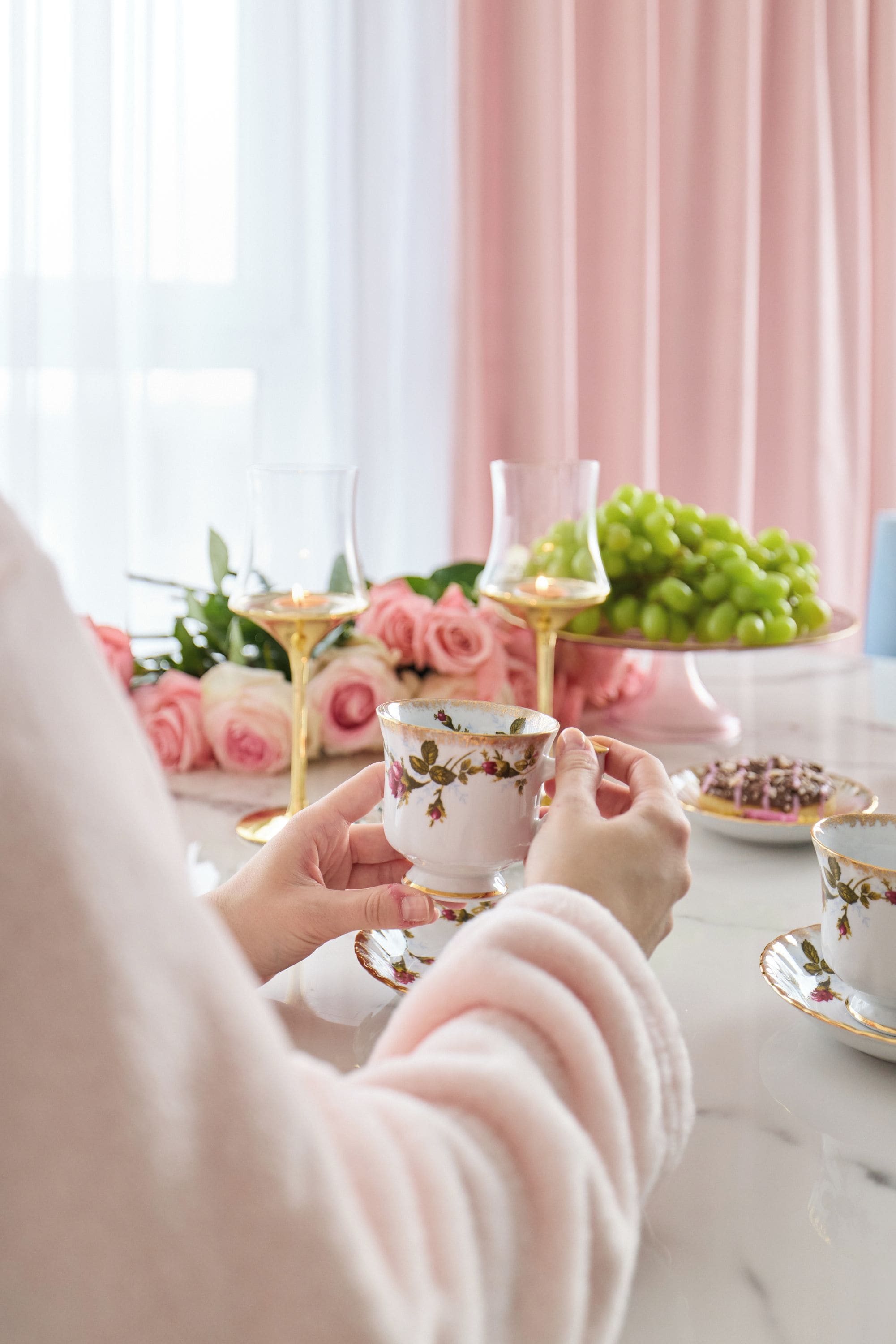 A person holding a teacup with floral designs, a glass of white wine, and a plate of green grapes.