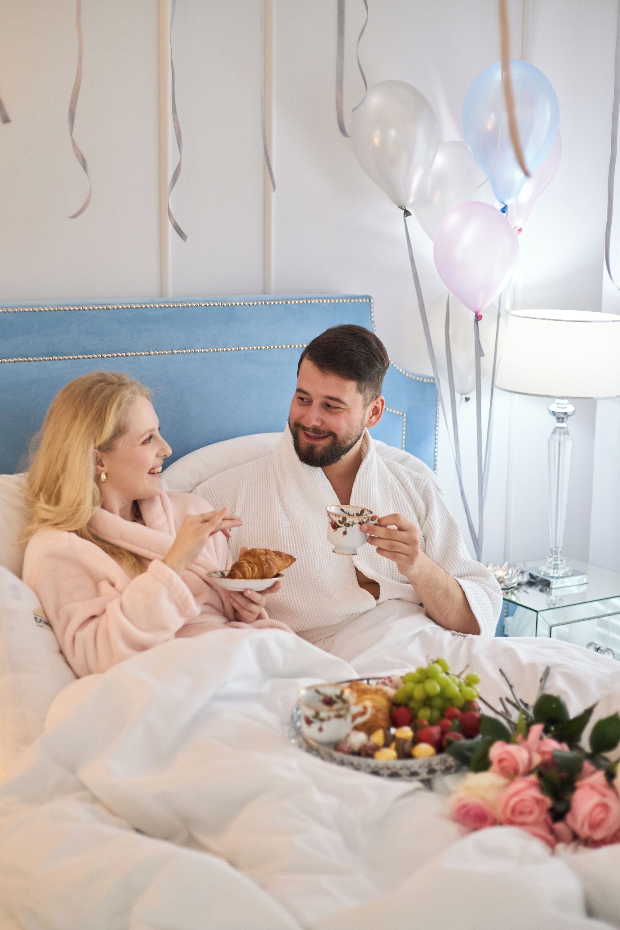 A couple in a bedroom with a bed, a bedside table with a lamp, and balloons in the background.