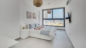 A modern bedroom with a large window, a white bed, a nightstand, and a wall-mounted TV.