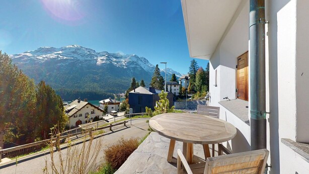 Een balkon met een houten tafel en stoelen met uitzicht op een berglandschap met besneeuwde toppen.