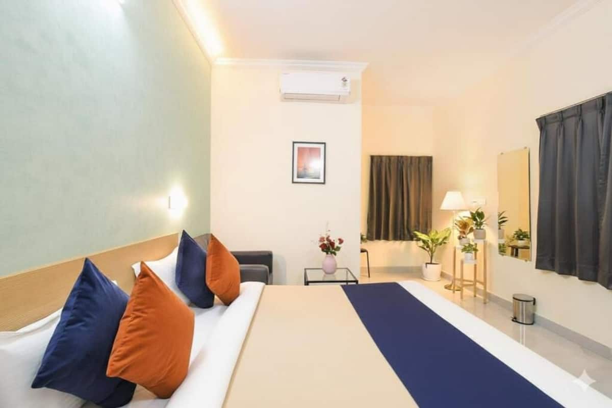 Habitación de hotel con cama, mesita de noche con lámpara, una pequeña mesa con un florero y una ventana con cortinas.
