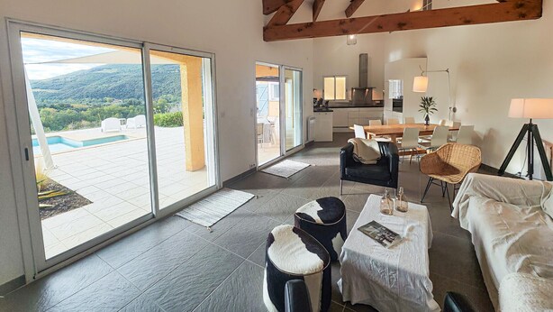 Un vaste espace de vie comprenant un grand salon avec une grande porte-fenêtre coulissante donnant sur un balcon, un coin repas avec une table et des chaises, et une cuisine située au fond de la pièce.