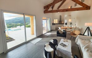 Un vaste espace de vie comprenant un grand salon avec une grande porte-fenêtre coulissante donnant sur un balcon, un coin repas avec une table et des chaises, et une cuisine située au fond de la pièce.