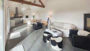 Un salon moderne comprenant un canapé, deux fauteuils, une table basse et une table à manger avec des chaises. On aperçoit une cuisine en arrière-plan, avec une cuisinière et des placards.