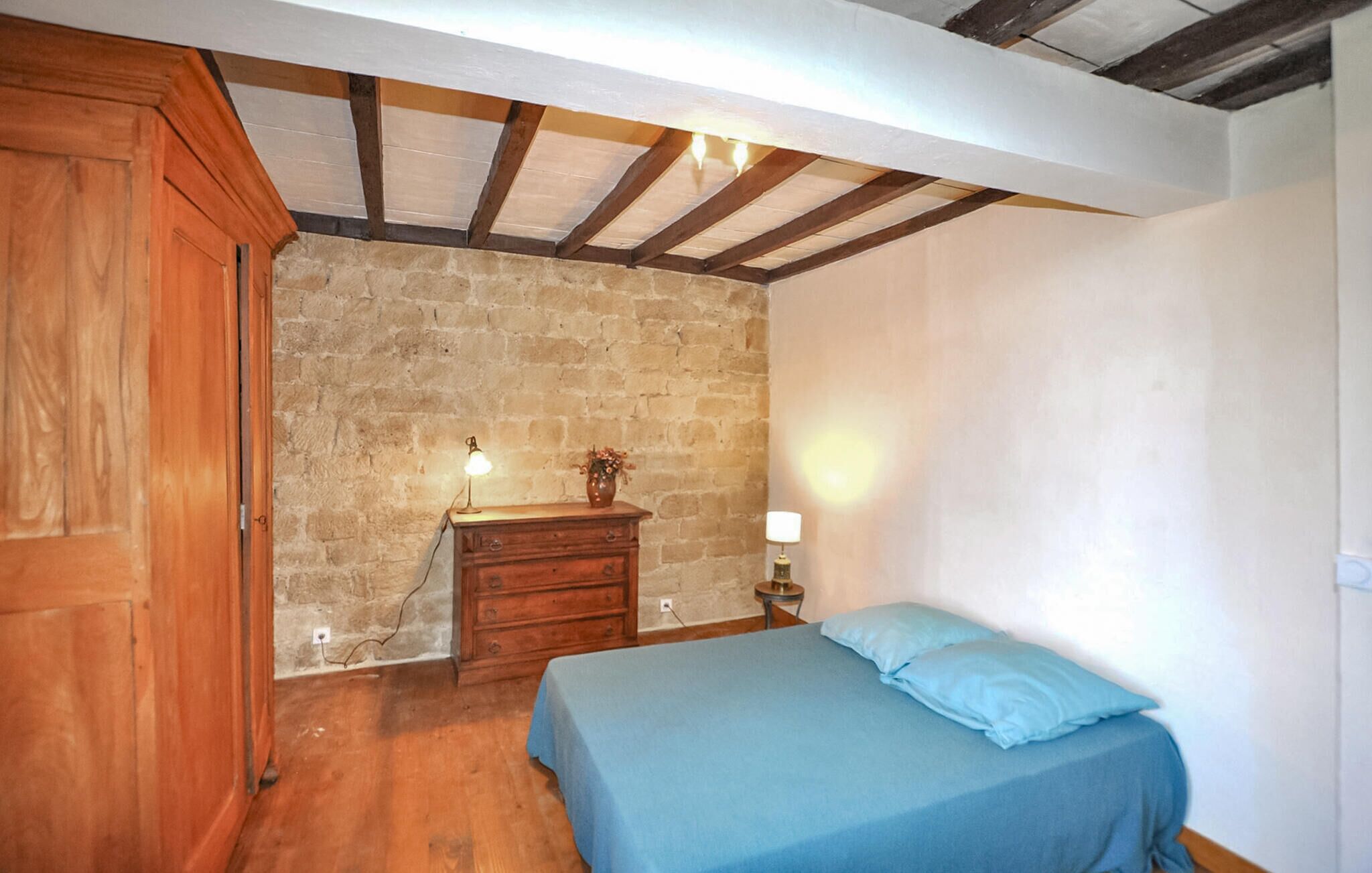 A bedroom with a stone wall, wooden ceiling beams, a wooden wardrobe, a dresser, a bedside table with a lamp, and a bed with blue bedding.