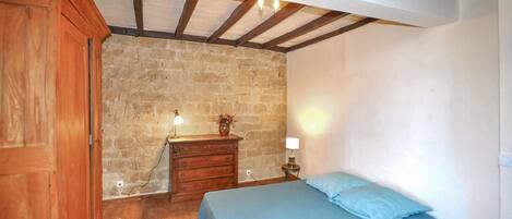 A bedroom with a stone wall, wooden ceiling beams, a wooden wardrobe, a dresser, a bedside table with a lamp, and a bed with blue bedding.