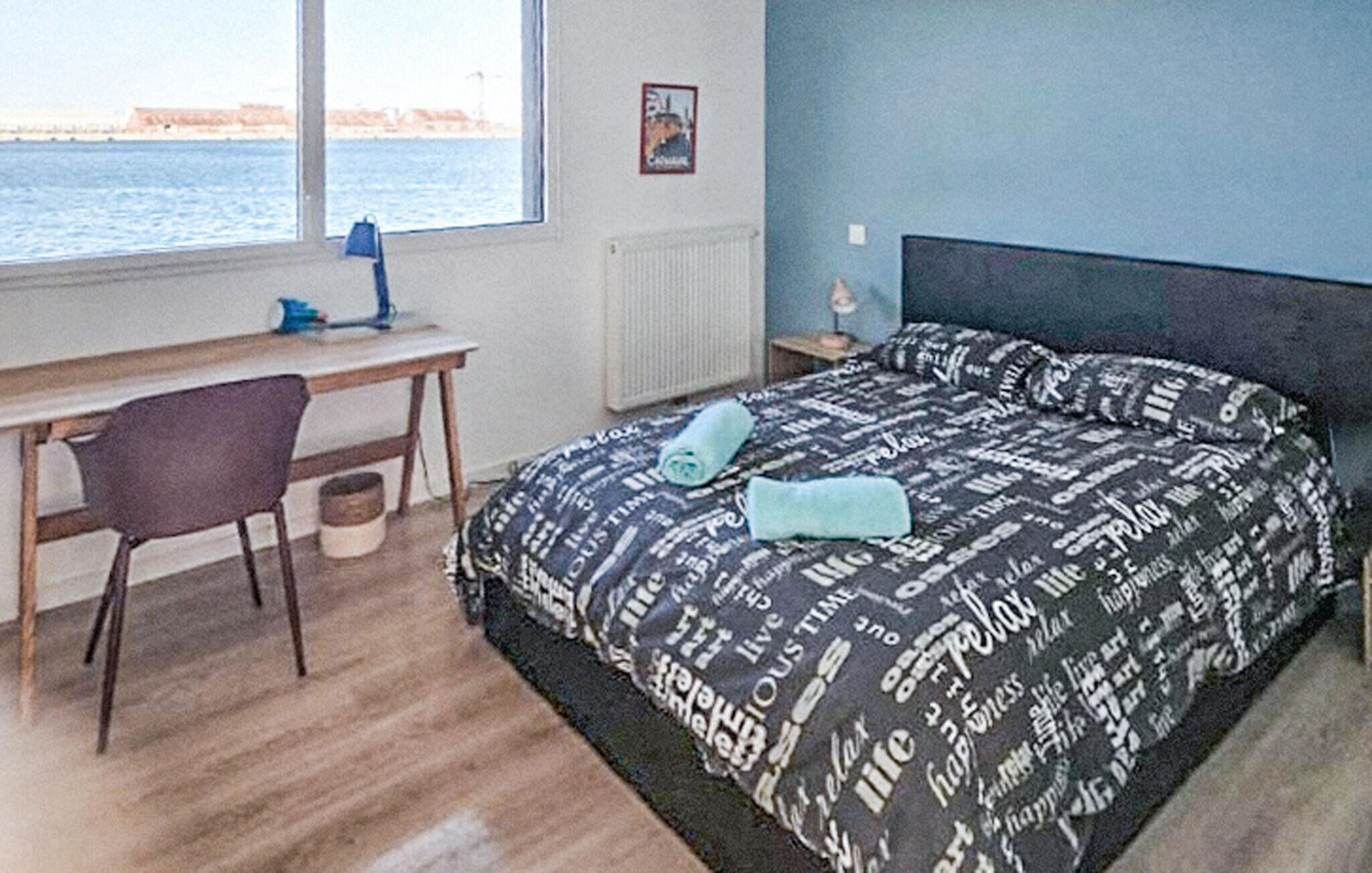 A bedroom with a bed, a desk, and a chair. There is a window with a view of the sea.