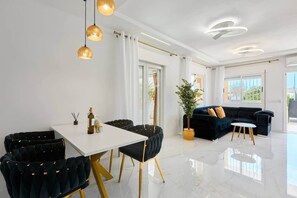 Una sala moderna con una mesa de comedor blanca, sillas tapizadas oscuras, un sofá oscuro y una planta en maceta.