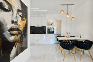 Une cuisine moderne avec des armoires blanches, un comptoir noir et des éléments décoratifs dorés. On y trouve un coin repas avec une table blanche et des chaises foncées.
