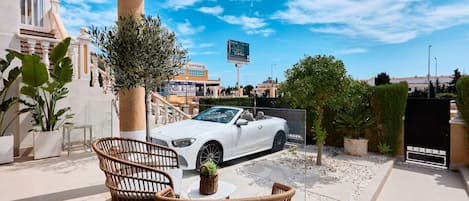 Une voiture décapotable blanche est garée dans une allée, avec un ciel dégagé au-dessus. On aperçoit un bâtiment blanc doté d’un balcon, ainsi que du mobilier d’extérieur.