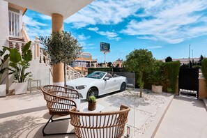 Ein weißer Cabrio parkt auf einer Auffahrt, der Himmel ist wolkenlos. Im Hintergrund ein weißes Gebäude mit Balkon und Gartenmöbeln.