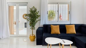 Een moderne woonkamer met een donkerblauwe bank, twee witte ronde tafels, een potplant en uitzicht naar buiten door een raam met metalen tralies.