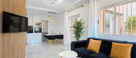 A modern living room with a black sofa, two white side tables, and a flat-screen TV mounted on a wooden panel.
