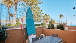 An outdoor seating area with a table and chairs, a surfboard leaning against a wall, and a view of the ocean. - Conquistador C (El Campello)