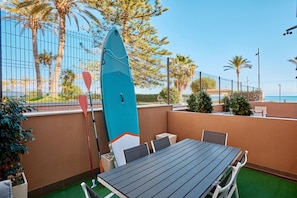 An outdoor seating area with a table and chairs, a surfboard leaning against a wall, and a view of the ocean.