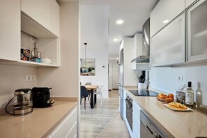 A modern kitchen with white cabinetry, a coffee machine, a kettle, and a dining area with a table and chairs. - Conquistador C (El Campello)