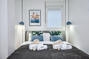 Un lit bien fait, recouvert d’une couvre-lit à rayures, avec deux oreillers aux motifs verts et blancs, et deux serviettes enroulées, portant un logo.