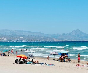 A beach with people sunbathing and swimming, umbrellas, and a mountain in the background.