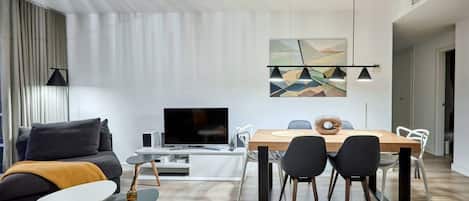 A modern living room with a flat-screen TV, a dining area with a wooden table and chairs, and a large abstract painting on the wall.