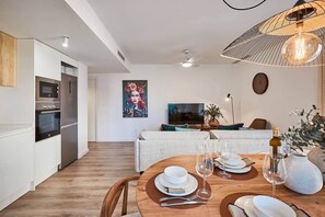 A modern kitchen with built-in appliances, a dining area with a round table set for four, and a living space with a sofa and television.