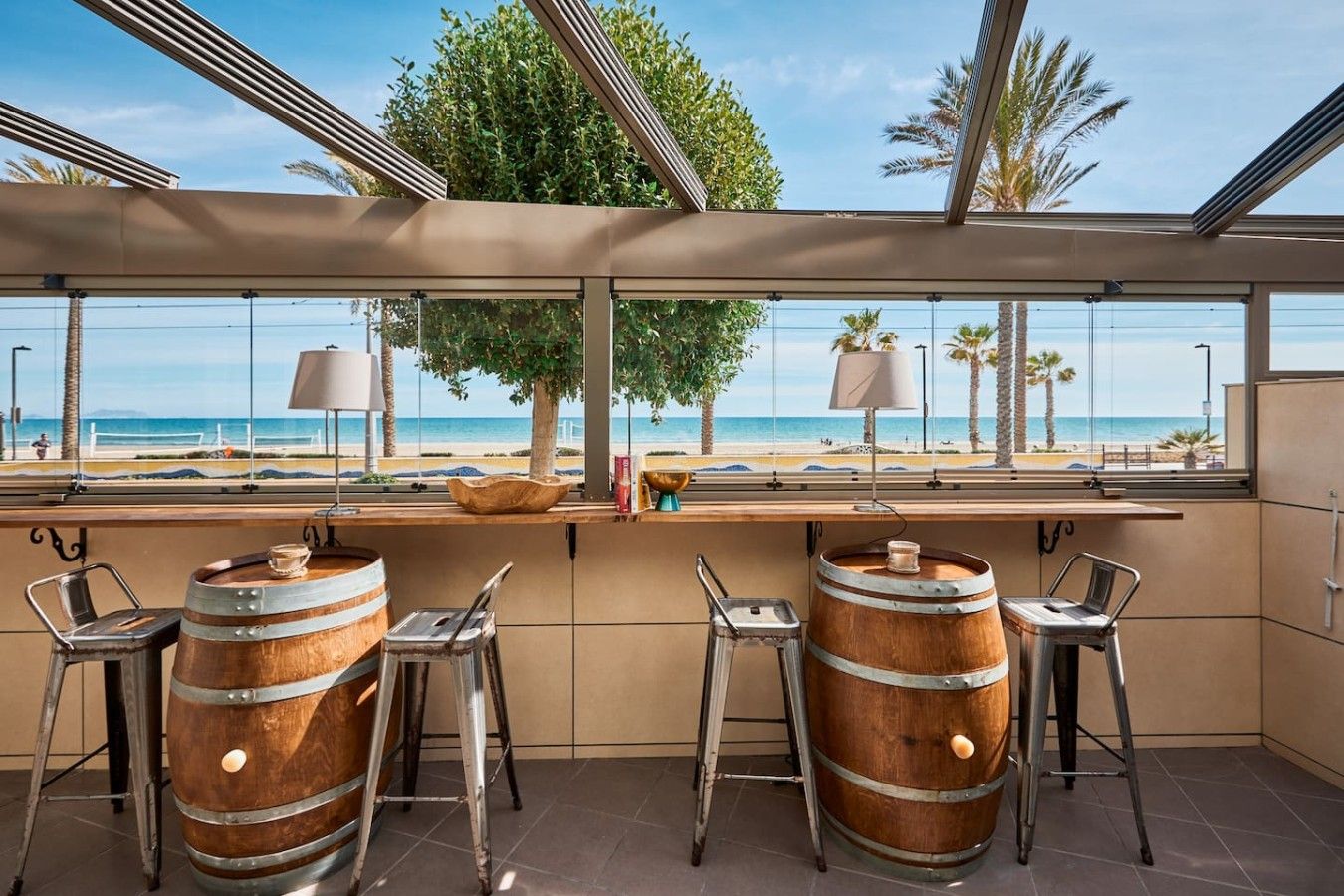 Un espace bar avec vue sur la plage, agrémenté de barils en bois, de tabourets en métal et d’une grande fenêtre.