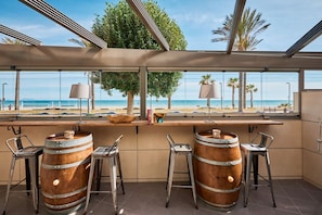 A bar area with a view of the beach, featuring wooden barrels, metal stools, and a large window.