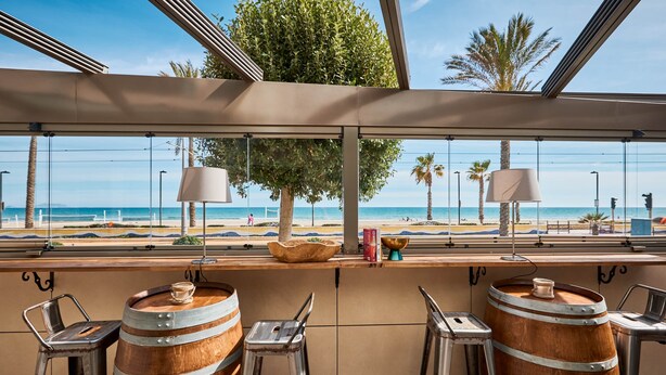 Uma área de bar com barris de madeira, banquetas de metal e vista para a praia e palmeiras.