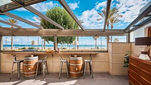 Área de bar com grandes janelas de frente para uma praia, com barris de madeira e banquetas de metal.