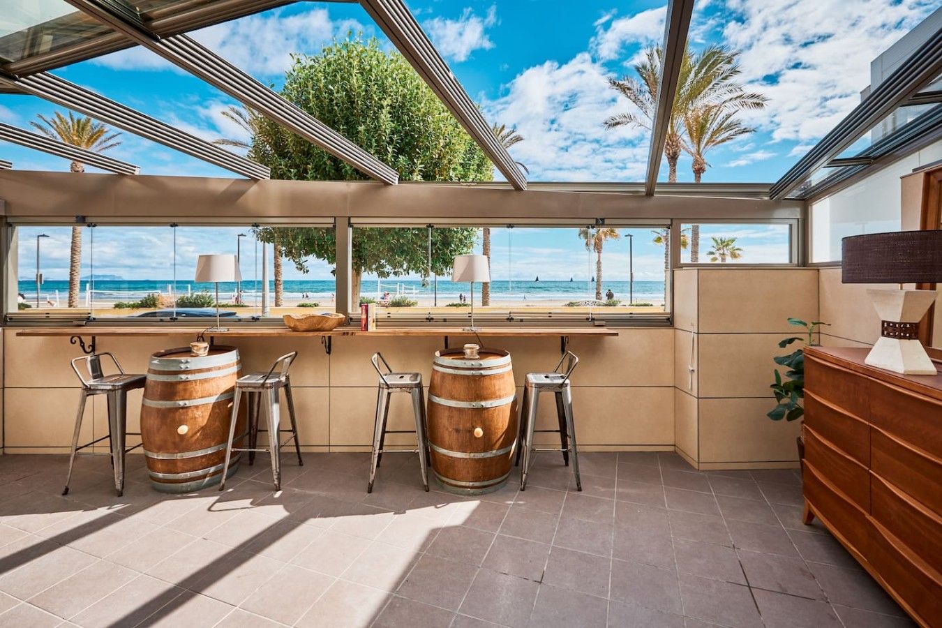 A bar area with wooden barrels, metal stools, and a view of the beach.