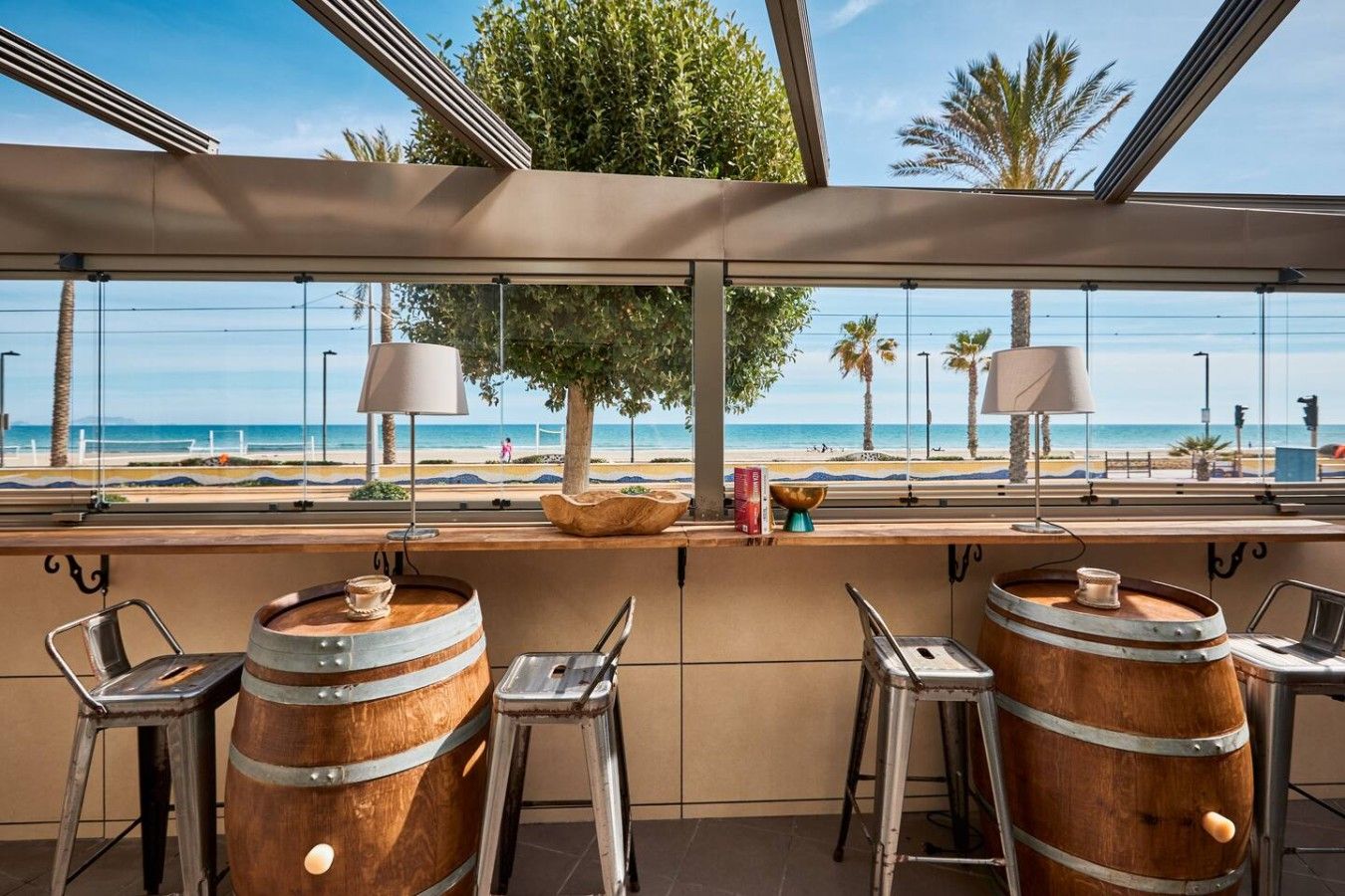 A bar area with wooden barrels, metal stools, and a view of the beach.