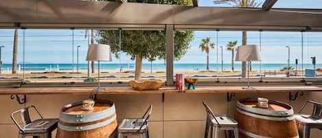 A bar area with wooden barrels, metal stools, and a view of the beach.