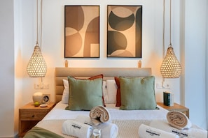 Een hotelkamer met een bed, twee aan de muur bevestigde abstracte schilderijen, twee hangende pendantlampen, een houten nachtkastje met een klein tafeltje, en opgerolde handdoeken met 'Welcome'-branding.