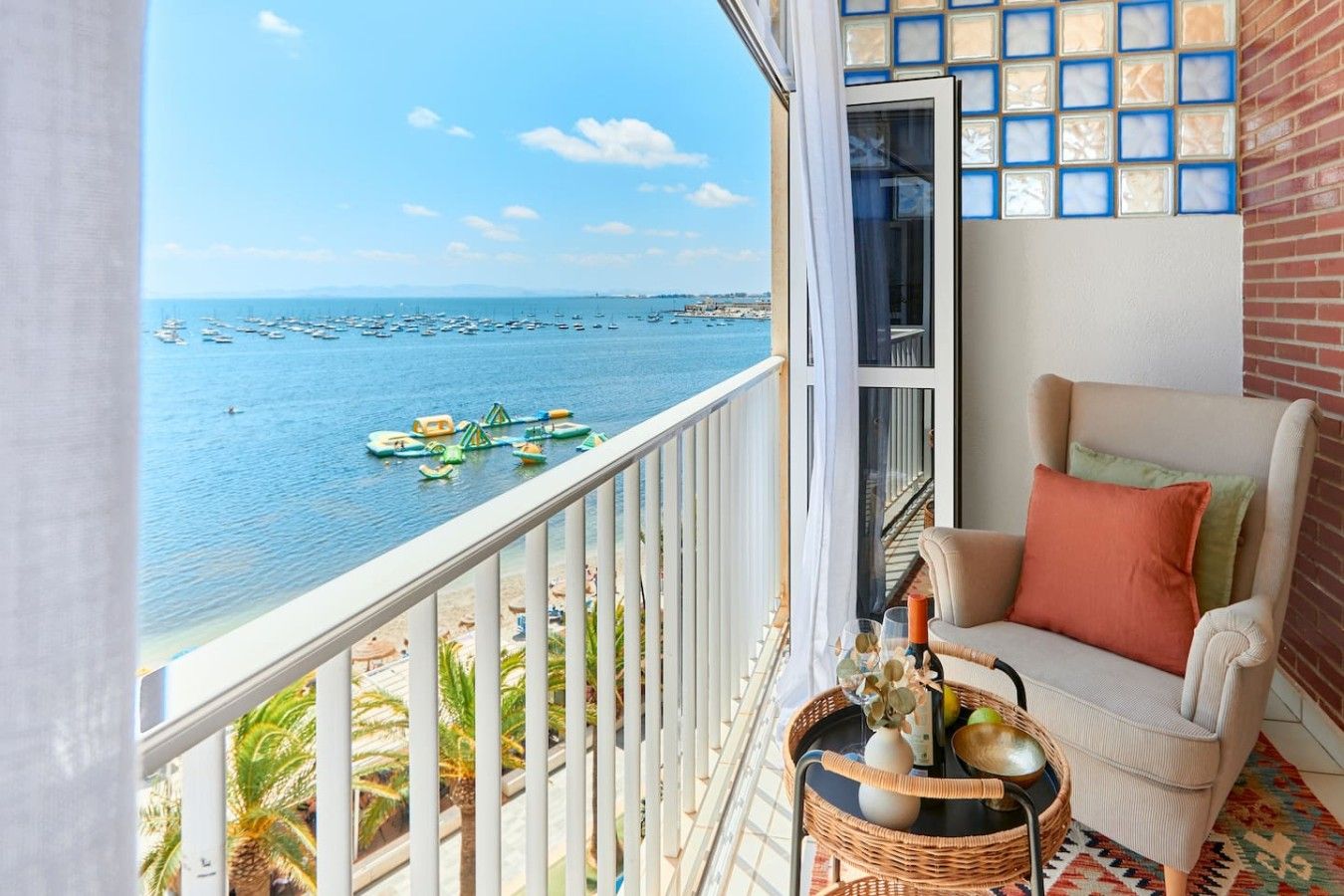 A balcony with a view of the sea, a wicker chair with cushions, and a small table with a vase and a bottle.