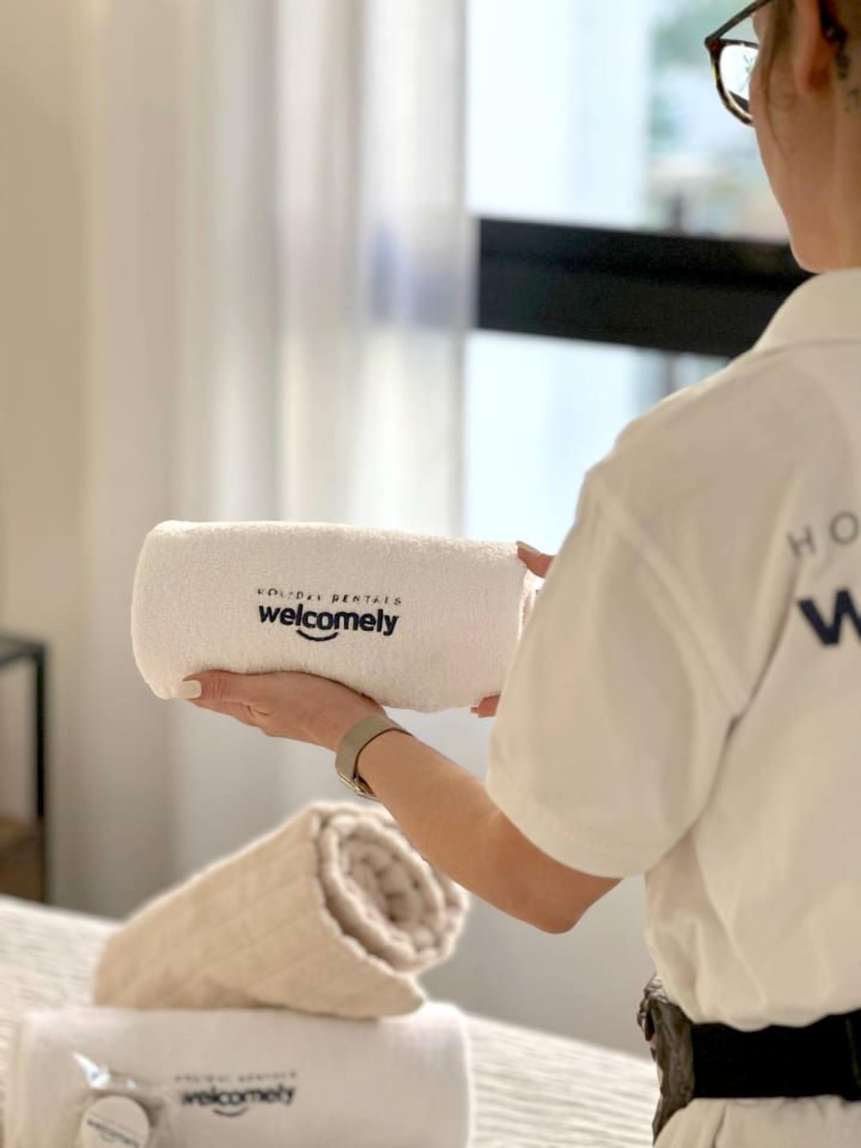 Eine Person hält ein gerolltes Handtuch mit 'welcomely'-Branding in der Hand, stehend in einem Zimmer mit Bett und Fenster.