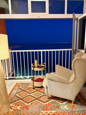 A balcony with a view of the sea at night, a wicker side table with a book, a patterned rug, and a beige armchair.
