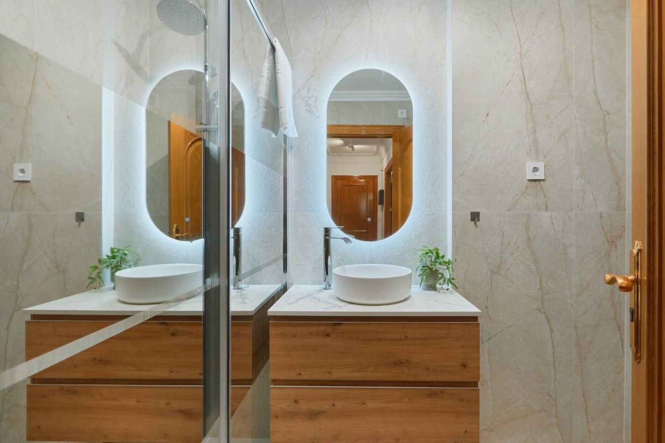 A bathroom with two sinks, wooden vanity, marble walls, and a glass shower enclosure.