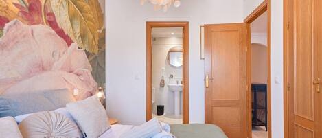 A hotel room with a bed, a large floral tapestry, a wooden door, and a bathroom visible through an open door.