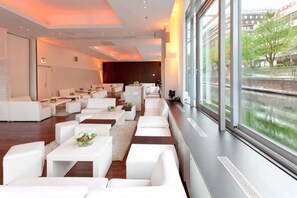 Un moderno hall de hotel con sofás blancos, mesas y un amplio ventanal con vistas a un río.