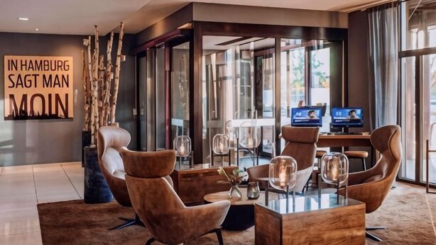 A modern hotel lobby with a glass partition, leather armchairs, a wooden coffee table, and a large digital screen.