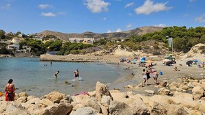 Ein felsiger Strand mit klarem Wasser, Menschen beim Schwimmen und Sonnenbaden, und Gebäude im Hintergrund.