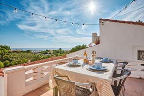 Un área de comedor al aire libre con una mesa dispuesta para dos personas, rodeada de sillas de mimbre, bajo luces colgantes, con vista al mar y follaje en el fondo.