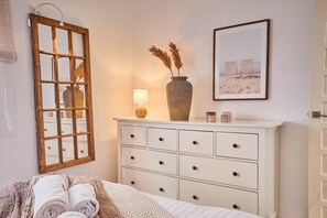 Un dormitorio ordenado con cama, cómoda con cajones, espejo, lámpara, florero con plantas secas y un cuadro en la pared.